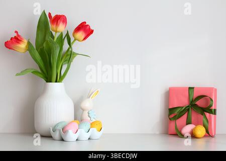 Vase of tulips, Easter bunny, colorful eggs, gift box on the table against the white wall. Stock Photo