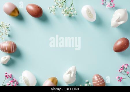 Happy Easter greeting card mockup. Frame of Easter eggs, minimalist flowers, decorative bunnies on pastel blue background. Flat lay, top view. Stock Photo