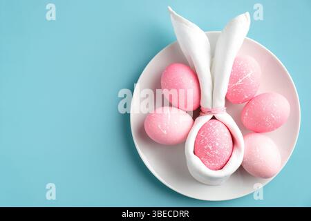 Stylish Easter table setting with easter bunny napkin and pink eggs on plate on turquoise background. Flat lay, top view. Stock Photo