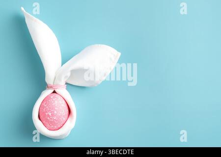 Easter bunny rabbit with funny ears made of napkin and pink egg on turquoise background. Minimal style Easter greeting card template. Stock Photo