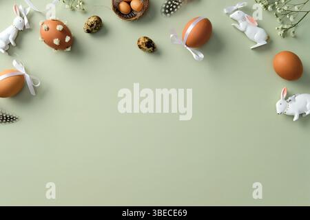 Easter flat lay composition with decorated eggs, ceramic bunnies, feathers, and flowers on a pastel green background. Stock Photo
