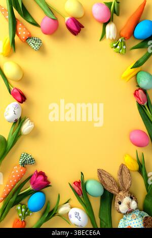 Frame made of painted Easter eggs, festive decorations, and tulip flowers on yellow background. Stock Photo