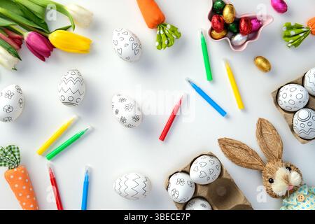 Easter flat lay composition with decorated eggs, colorful markers, tulips, and bunny toy on a white background. DIY, handicraft Easter eggs decoration Stock Photo
