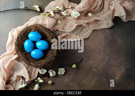 Nest with four vibrant blue Easter eggs on a rustic background, adorned with delicate white blossoms and soft pink fabric Stock Photo