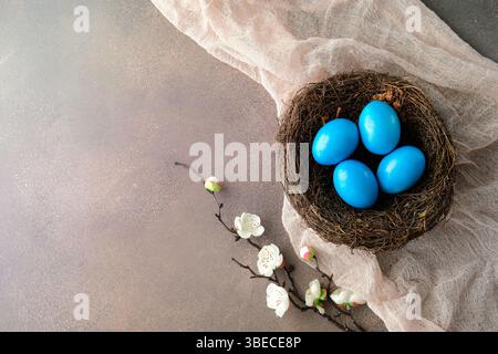 Vibrant blue bird eggs in a natural nest on soft fabric, adorned with delicate white blossoms. Happy Easter concept Stock Photo