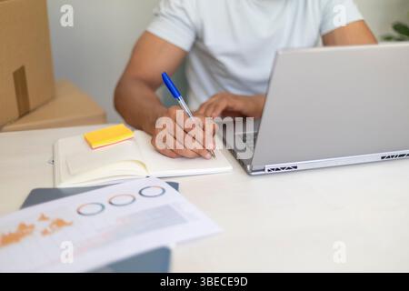 close up of a focused man multitasking with a laptop and notebook, running a local business Stock Photo