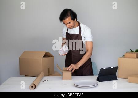 Man wrapping an item in bubble wrap and placing it into a small box, preparing packages on a table with boxes and plates Stock Photo