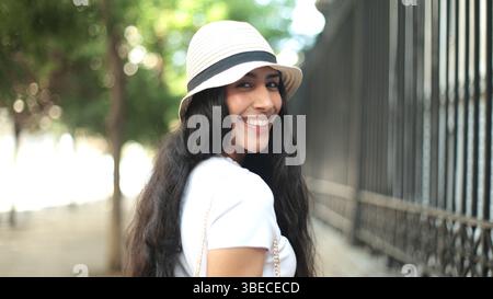 Close-up shot of a young latin woman with long hair wearing a hat and white top, smiling brightly. The outdoor background is softly blurred, emphasizi Stock Photo