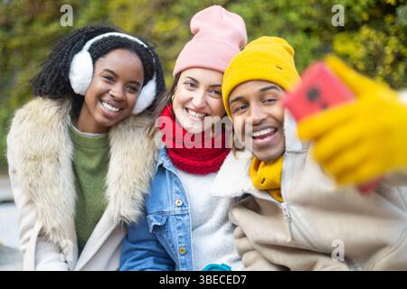 Three joyful young friends bundled in winter clothes, smiling and taking a selfie together in a chilly park, enjoying their time outdoors Stock Photo
