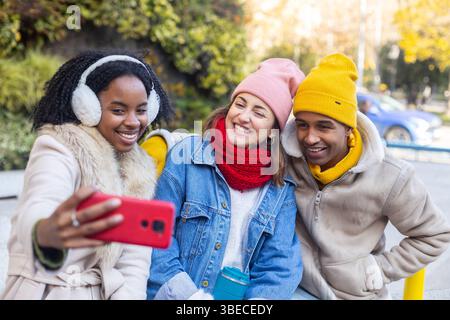 Three cheerful young friends wearing winter clothes and colorful beanies taking a selfie outdoors with a smartphone Stock Photo