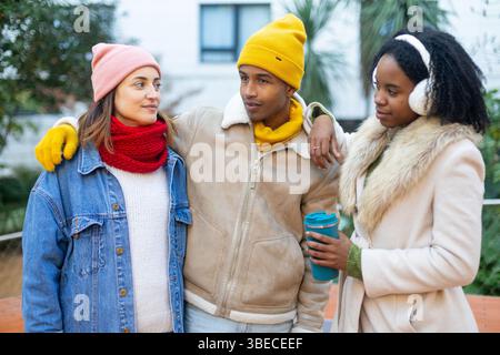 Three cheerful young multi ethnic friends wearing winter clothes, walking together outdoors and enjoying a hot drink Stock Photo
