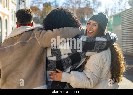 Three young friends walking down a city street, enjoying a sunny winter day and hugging each other Stock Photo
