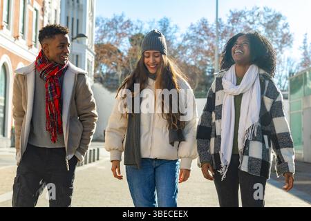 Three cheerful multi ethnic young students walking and chatting together on a sunny winter day on campus Stock Photo