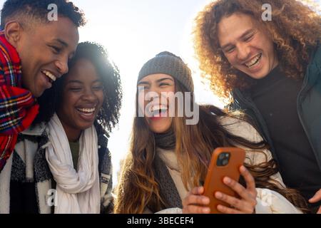 Four cheerful young friends are having fun together outdoors in winter, sharing funny content on a smartphone Stock Photo