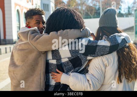Three university students are walking together embracing each other on a sunny winter day Stock Photo