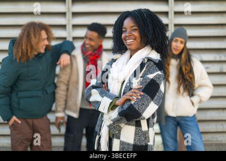 Confident young woman with crossed arms stands in front of her friends, smiling on a cold winter day Stock Photo