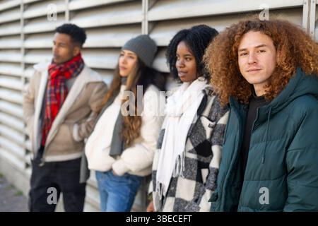 Four young people dressed in trendy winter clothes are standing together against a metal wall, showcasing their urban style and diversity Stock Photo