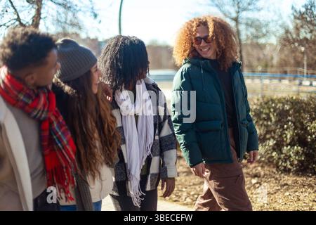 Four cheerful multi ethnic young students are walking and chatting together in a park during a sunny winter day Stock Photo