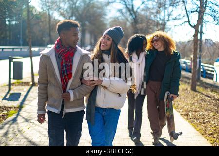Happy multi ethnic young couple strolling arm in arm on sunny winter day in city park, enjoying time together with friends Stock Photo