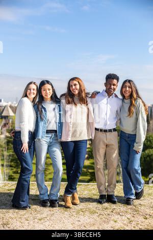 Five multi ethnic students standing together in a park with arms around each other, smiling Stock Photo