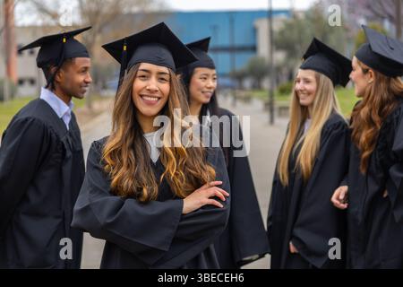 Portrait of cheerful graduate student smiling with arms crossed while standing with her colleagues outdoors after the graduation ceremony Stock Photo