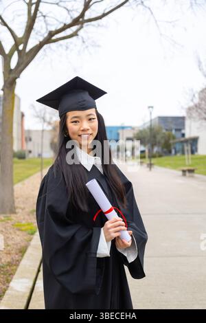 Young woman wearing graduation gown and cap proudly holding diploma, celebrating academic achievement on university campus Stock Photo