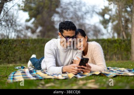 Young latin couple using mobile phone, lying on a blanket in a park, enjoying a sunny day Stock Photo