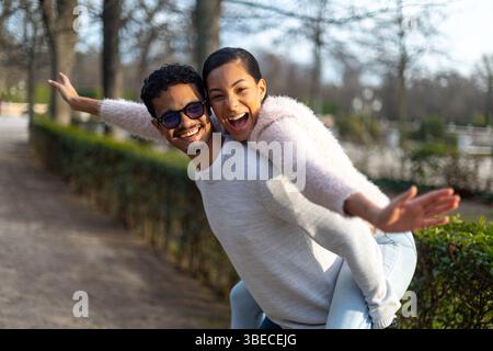 Young man giving piggyback ride to girlfriend in a park, both with arms outstretched and laughing Stock Photo