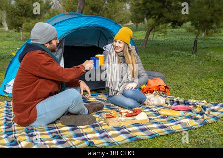 Young couple toasting with mugs while having a picnic in front of their tent during a camping trip Stock Photo