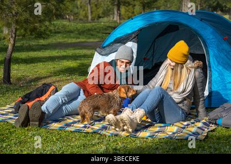Young couple enjoying a relaxing camping trip with their dachshund dog, sitting by their tent in a sunny park Stock Photo