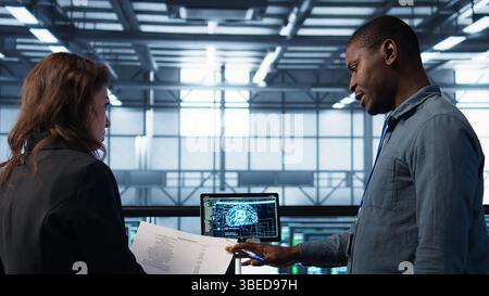 Team of IT professionals looking over data center checkup paperwork, using AI tech on tablet. Colleagues collaborating in server farm, reading documentation, using artificial intelligence, camera B Stock Photo