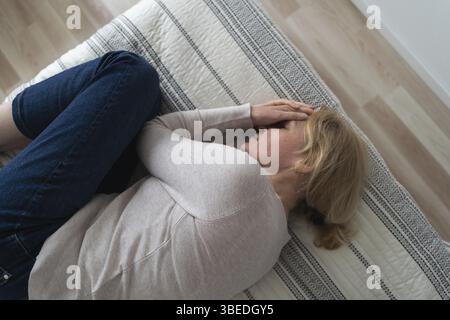 Mature woman curled lying on the bed at home top view. Depression, mental health, abuse problem. Stock Photo