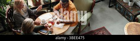Three people, seated around a table in cozy cafe, engaging in lively conversation while enjoying coffee. Green plants and comfortable furniture create a relaxing ambiance Stock Photo