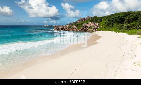 Waves washing the shore of a secluded beach with rocky cliffs and lush greenery. Grand Anse Beach, Seychelles.. Stock Photo
