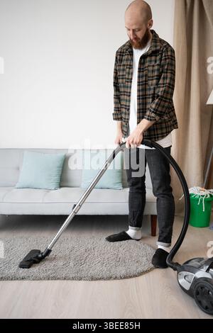 Cleaning and housekeeping concept. Man vacuuming carpet with vacuum cleaner during daily housework at home interior. Stock Photo