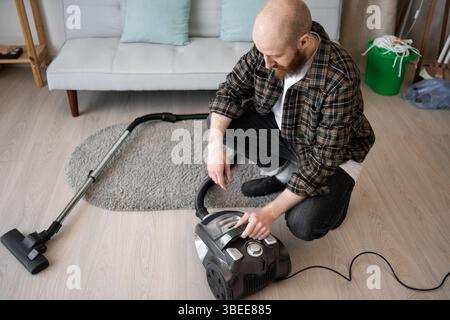 Young handsome man with beard cleaning house sitting near vacuum cleaner starts vacuuming Stock Photo