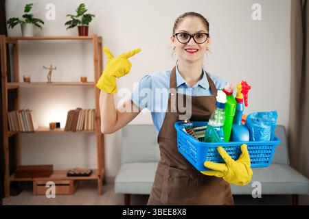 Cleaner woman wearing apron holding cleaning products in basket standing in the living room pointing with fingers teeth. cleaning concept Stock Photo