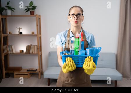 Female cleaner in apron and yellow gloves with cleanser bottles in basket pretends to be kissing doing fish face. copy space Stock Photo