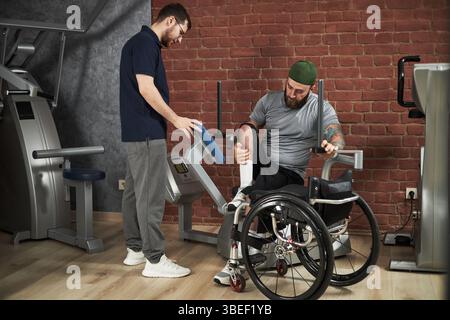 Trainer assisting disabled man in wheelchair exercising in gym. Man using gym equipment, wearing casual sports attire while trainer providing support Stock Photo