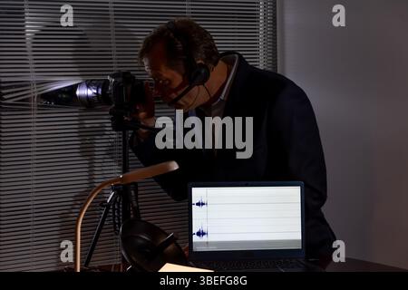 A private detective in a dark room with a camera and laptop records video and audio Stock Photo