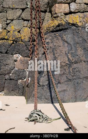 Chains hang from a stone wall onto a sandy beach, possibly for boat mooring, showing the effects of tide and time. Stock Photo