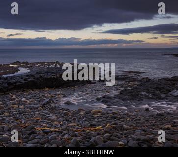 Dark basalt rocks and wet pebbles along a north western Scottish shoreline at dusk with moody skies and soft sea motion in winter. Stock Photo