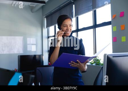 Late-twenties woman speaking on wireless headset and holding folder in office with monitors, prints Stock Photo