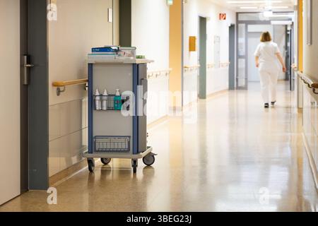 Deutschland 25. April 2025: Im Bild: Dieses Foto zeigt einen typischen, hellen und sehr sauberen Krankenhausflur in einer modernen medizinischen Einrichtung. Der glänzende Boden reflektiert das Licht, was auf hohe Hygienestandards und regelmäßige Reinigung schließen lässt. Entlang der Wände befinden sich Handläufe aus hellem Holz, die für Patienten und Besucher zusätzliche Unterstützung bieten. Im Vordergrund steht ein gut ausgestatteter, mobiler Pflege-Rollwagen, auf dem medizinische Utensilien und Desinfektionsmittel für den direkten Einsatz am Patienten bereitstehen. Dieser Wagen ist ein ze Stock Photo