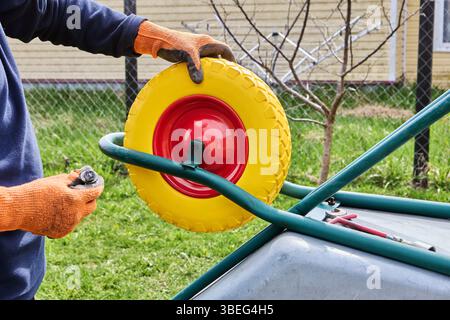 Holding replacement wheel and preparing axle for proper alignment before installation on garden wheelbarrow during outdoor maintenance task. Stock Photo