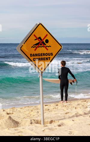 A surfer standing on the beach with a "Dangerous Current" sign in the foreground. Bronte Beach, Sydney, Australia Stock Photo