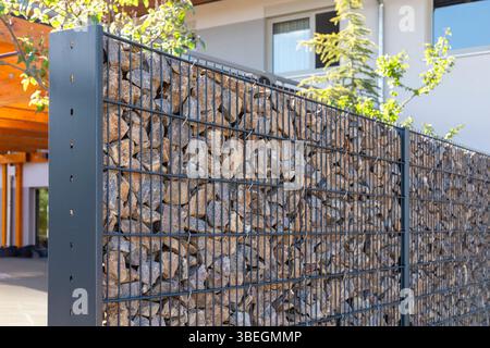 A modern gabion fence constructed from stones creates an elegant boundary. It stands in a contemporary setting, complemented by greenery and a sleek Stock Photo