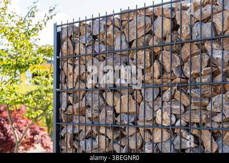 A modern gabion fence constructed from stones creates an elegant boundary. It stands in a contemporary setting, complemented by greenery and a sleek Stock Photo