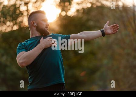 A sporty man listening to music on wireless headphones and doing stretching exercises before running or workout outdoor in the summer park active Stock Photo