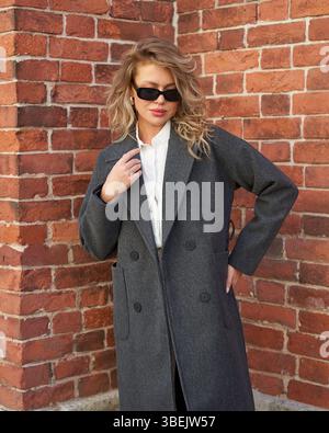 Blonde woman in stylish coat poses against red brick wall on a sunny city street Stock Photo
